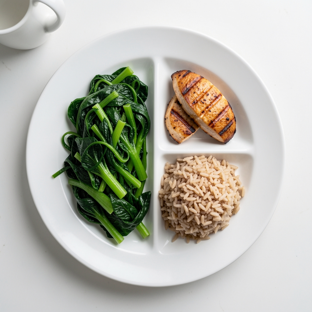 Top-down view of a white ceramic dinner plate divided into proportional zones with steamed green vegetables occupying half the plate, a small portion of grilled chicken fillet in one quarter, and brown rice in the remaining quarter on a clean white kitchen counter